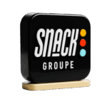 3D rendered black logo sign for Snack Groupe, featuring "SNACK" in bold white letters, "GROUPE" in smaller white letters below, and three colored dots (orange, red, light blue) vertically aligned on the right. The sign rests on a light beige base.