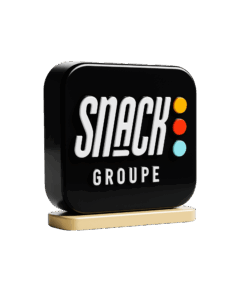 3D rendered black logo sign for Snack Groupe, featuring "SNACK" in bold white letters, "GROUPE" in smaller white letters below, and three colored dots (orange, red, light blue) vertically aligned on the right. The sign rests on a light beige base.